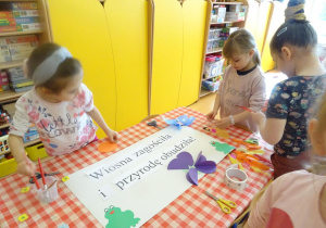 Trzy dziewczynka dekorują plakat, naklejają kolorowe kwiatki z różnego materiału plastycznego.
